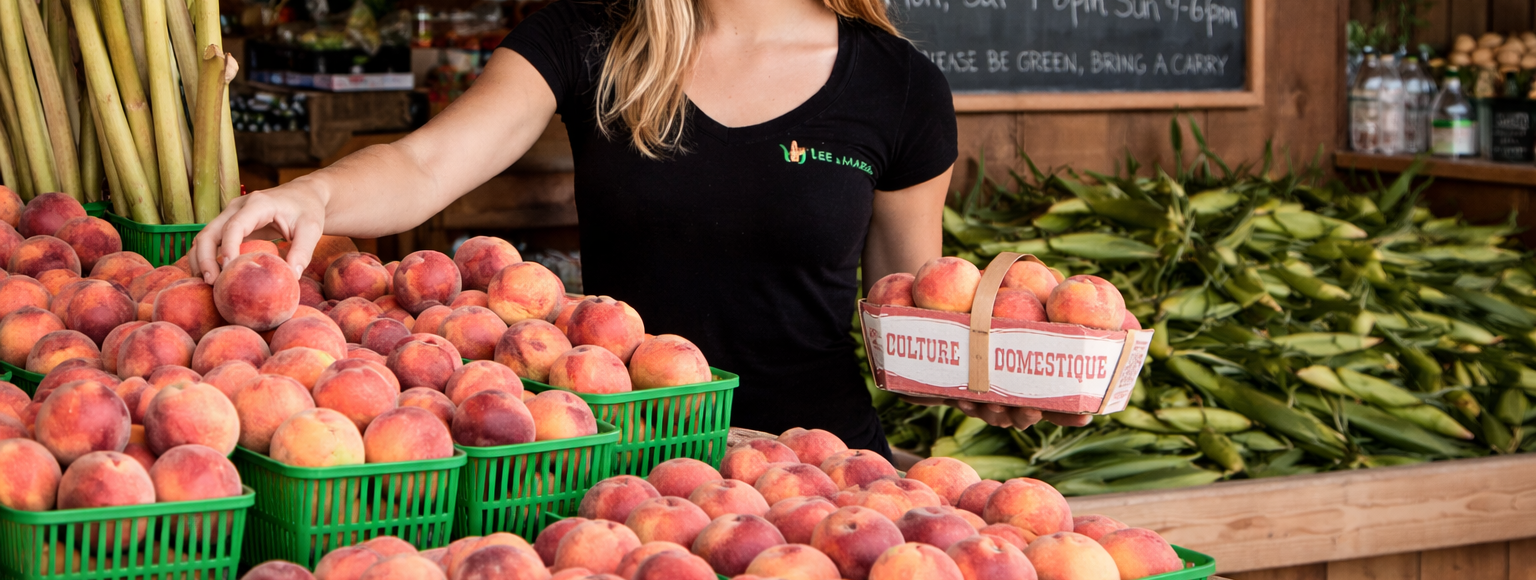 Woman arranging peaches at a farmers market with a chalkboard sign in the background.